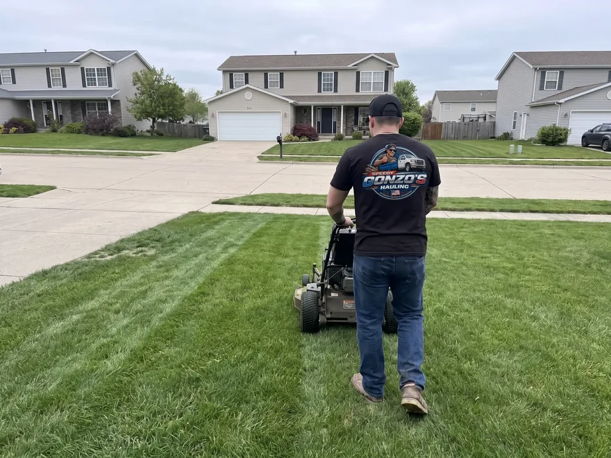 A Speedy Gonzo's Hauling crew member in a company t-shirt pushing a walk-behind mower across a suburban front lawn, fresh cut stripes in the grass behind him