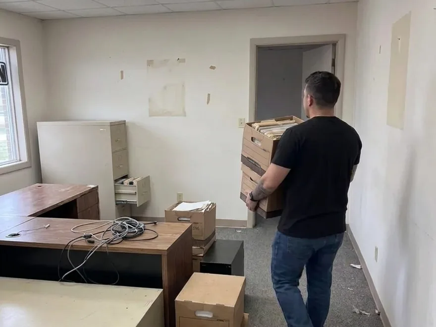An office being cleared out - a worker carrying stacked boxes of old files toward the door, past an emptied desk and an open filing cabinet