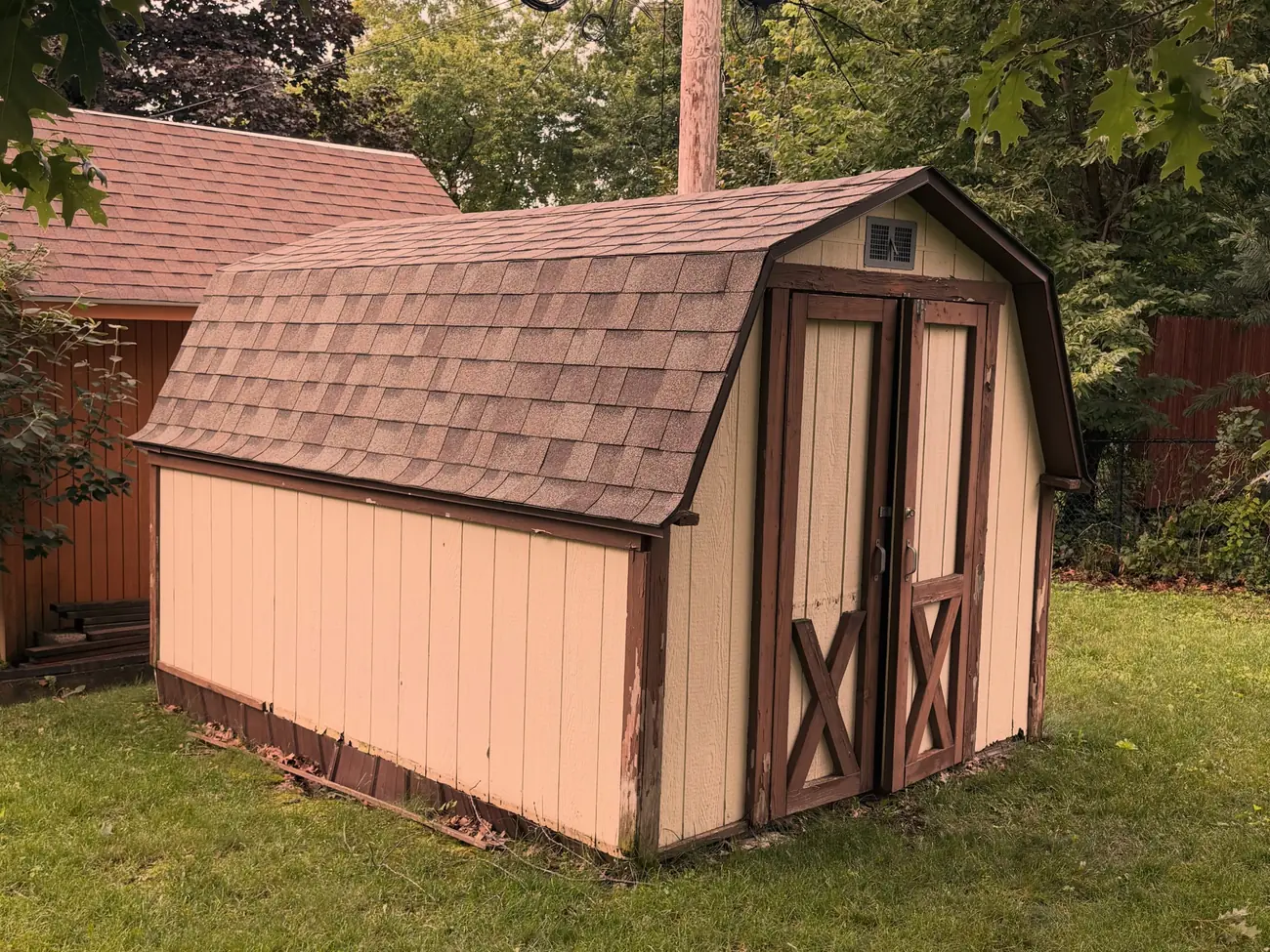 Tan gambrel-roof shed standing intact on the grass beside a red garage in a Peoria area back yard, before teardown