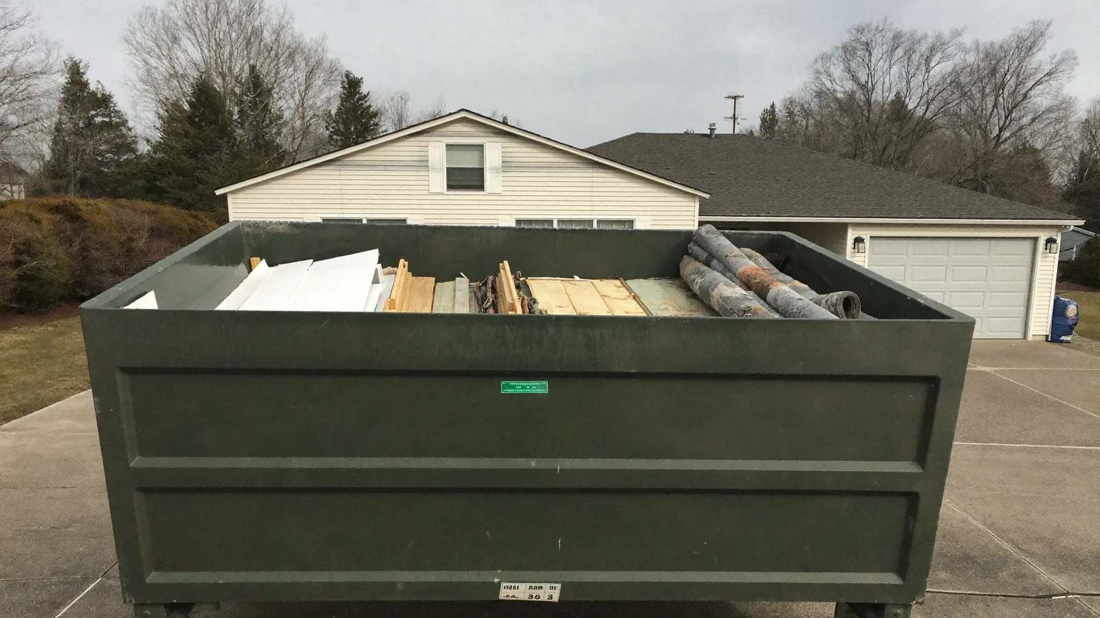 A plain green roll-off dumpster on a residential driveway, loaded with drywall sheets, lumber, and rolled carpet